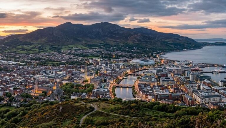 Aerial view of a coastal city at sunset with a river, bridges, and a mountain range in the background.