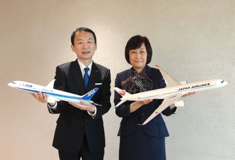 Two professionals in suits hold model airplanes, an ANA jet and a Japan Airlines jet, facing the camera in a neutral room.
