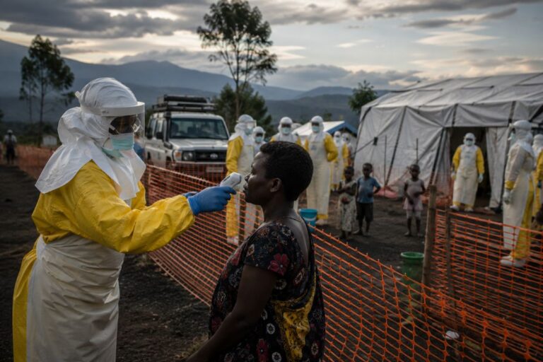 Healthcare worker in full PPE uses a handheld thermometer on a woman at a field clinic.