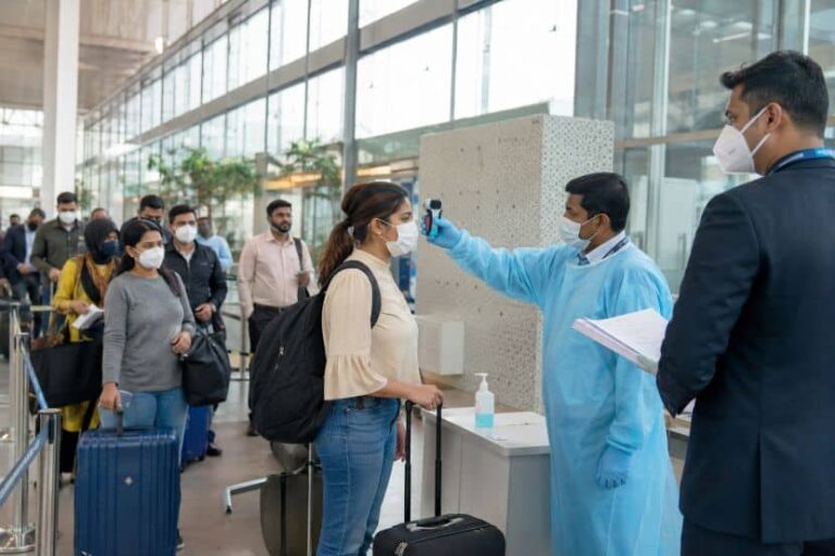 Travelers wearing masks line up at a health screening area as a staff member checks a passenger's temperature with a handheld thermometer.