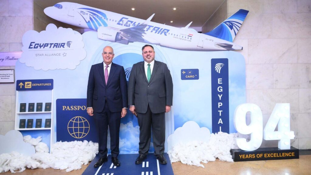 Two men in suits pose at an EgyptAir promotional display featuring a model plane and passport props for a 94th anniversary event.