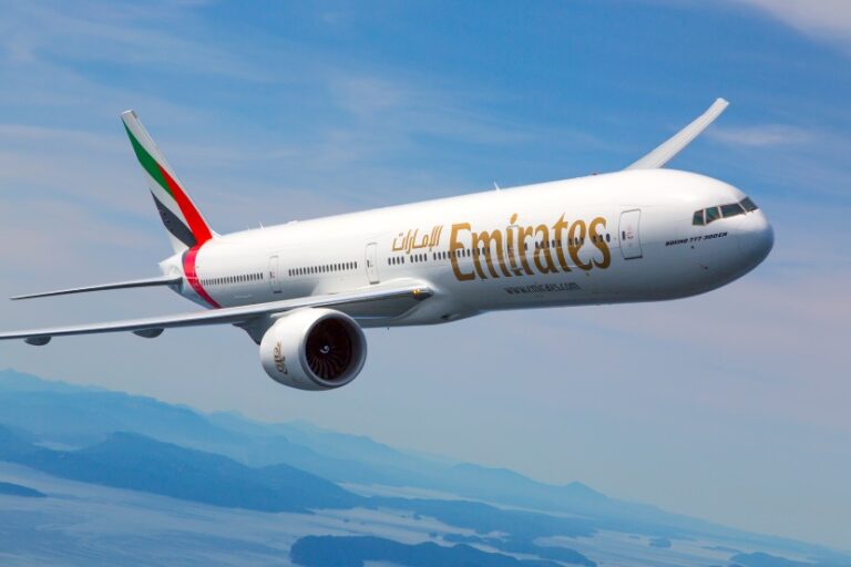 Emirates airliner in flight with blue sky and distant mountains below, tail decorated in Emirates colors.