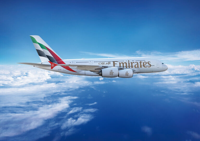 Emirates Airbus A380 flying above clouds against a blue sky with a white fuselage and large Emirates logo on the side, four engines visible.