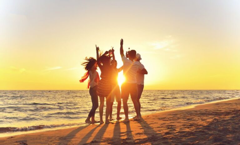 Group of friends on a sunlit beach, silhouetted against a golden sunset, raising their arms in celebration.