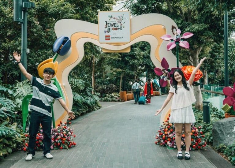 Two people pose with arms raised under a colorful arch at the Jewel Blooms botanical exhibit in a lush garden setting.