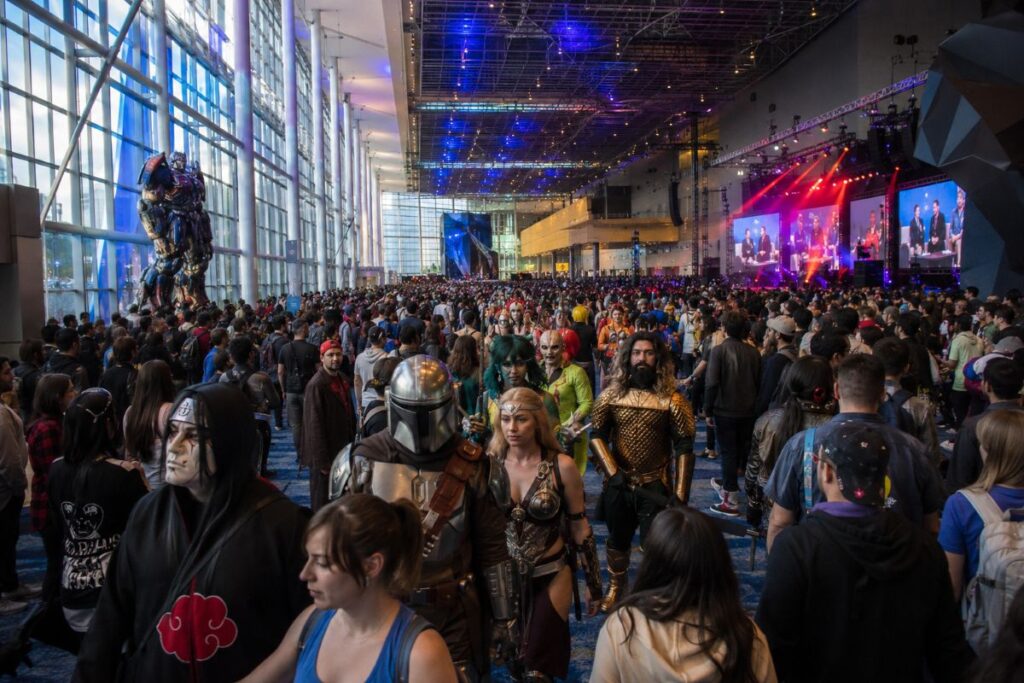 Large convention hall full of cosplayers and attendees with a stage and screens in the background, vibrant lighting throughout.