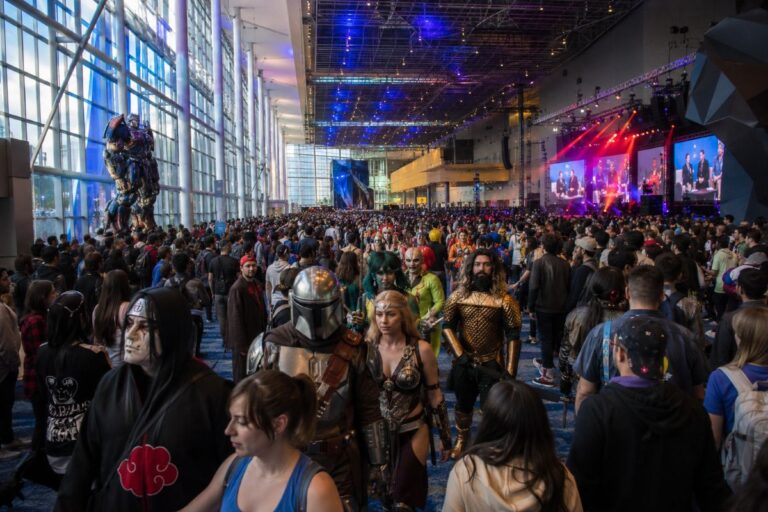 Large convention hall full of cosplayers and attendees with a stage and screens in the background, vibrant lighting throughout.