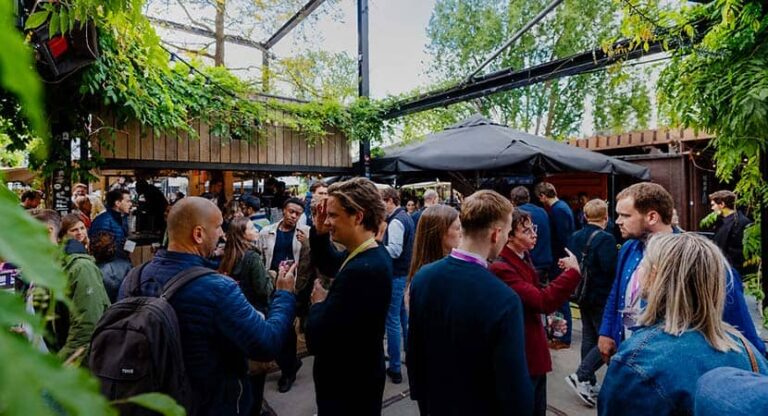 Group of professionals networking at an outdoor courtyard under a canopy, with greenery overhead and people chatting in small clusters.