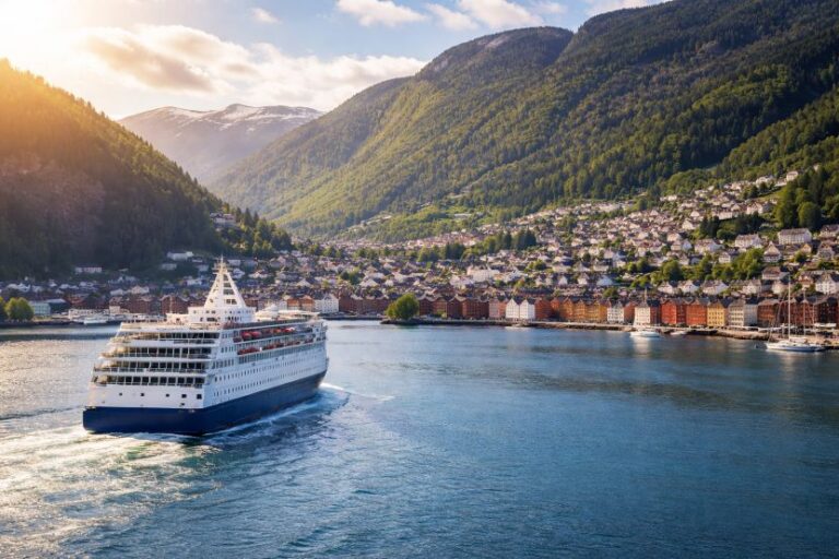 Cruise ship sailing past a coastal town with colorful buildings and green mountains in the background at sunset