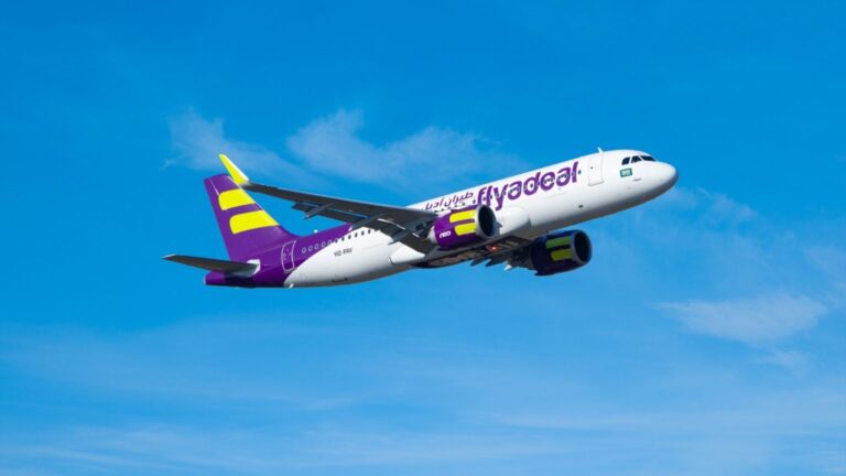 Airliner in flight with a purple and yellow tail, white fuselage, and Flyadeal branding against a clear blue sky.