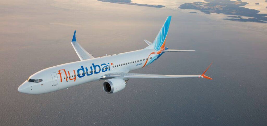 White Flydubai passenger jet flying over a calm sea with a sunset glow in the background, winglets orange and blue.