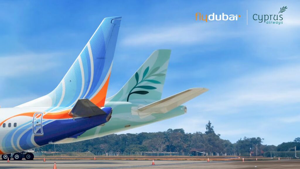 Rear view of two commercial aircraft tails on a runway, branded with flydubai and Cyprus Airways against a blue sky with orange cones nearby.