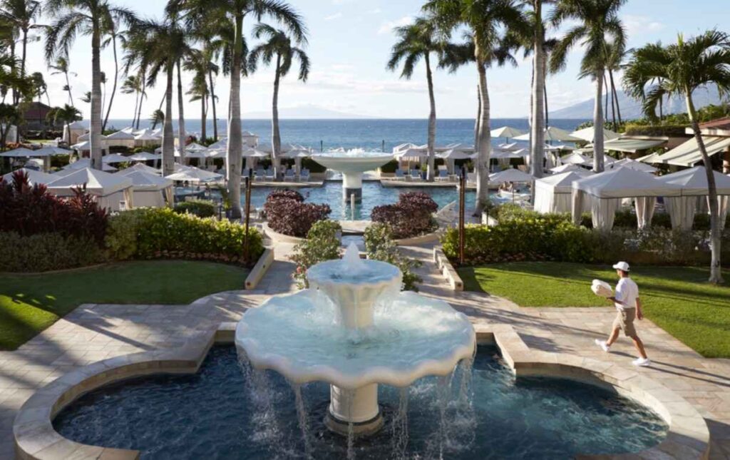 Resort pool with a large tiered fountain, white cabanas, and palm trees, ocean view in the background.