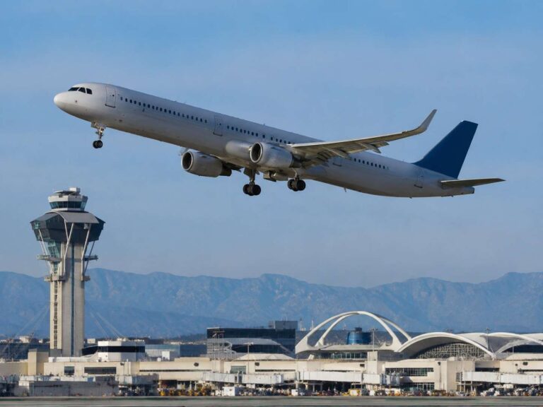 Airplane lifting off from the runway near an airport control tower with terminal buildings and distant mountains in the background.