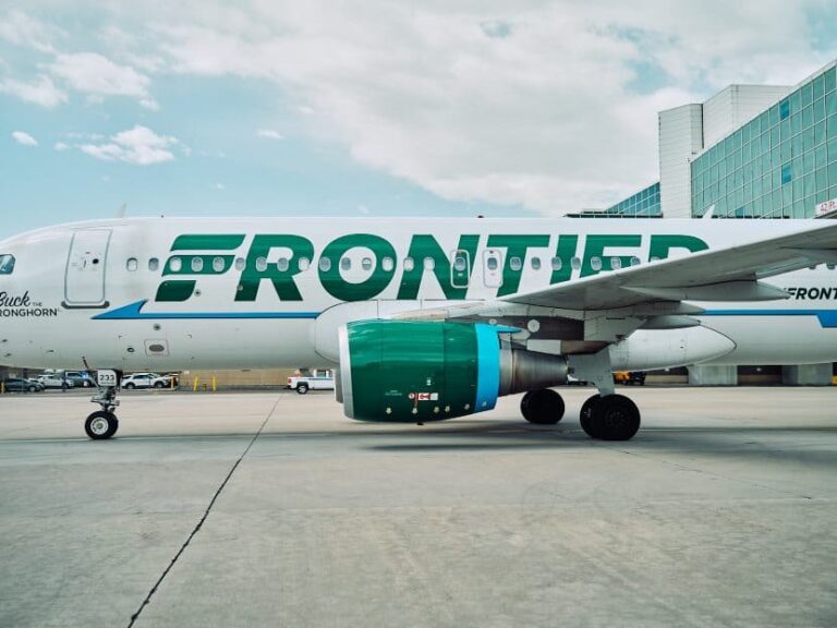 Frontier Airlines airplane on the tarmac beside a modern airport terminal, engine visible in the foreground.