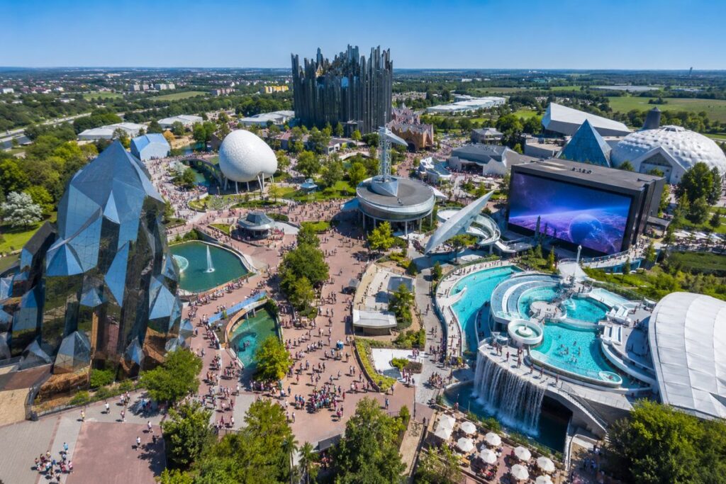 Aerial view of a futuristic theme-park with glass pyramids, a large white geodesic sphere, water features, and a giant outdoor screen filled with visitors wandering the paths.