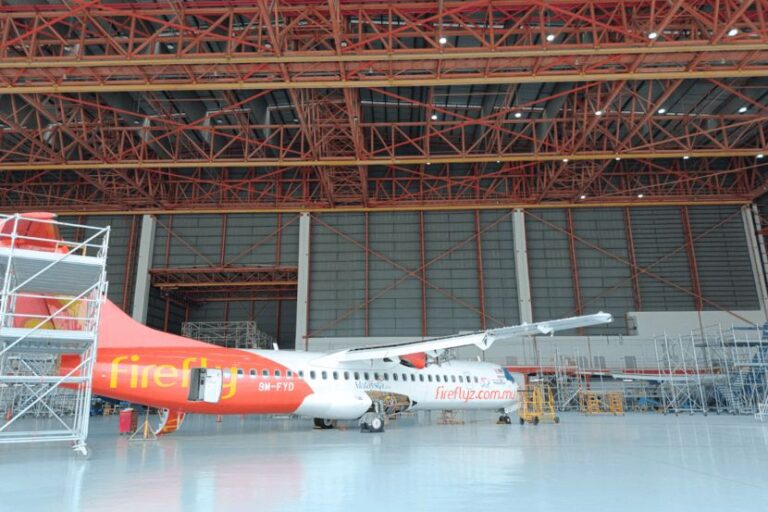 Red and white Firefly aircraft parked inside a spacious hangar with scaffolding and maintenance equipment nearby.