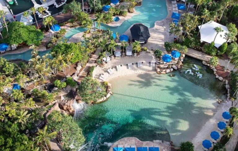 Aerial view of a tropical resort pool area with turquoise water, palm trees, lounge chairs, and blue umbrellas.