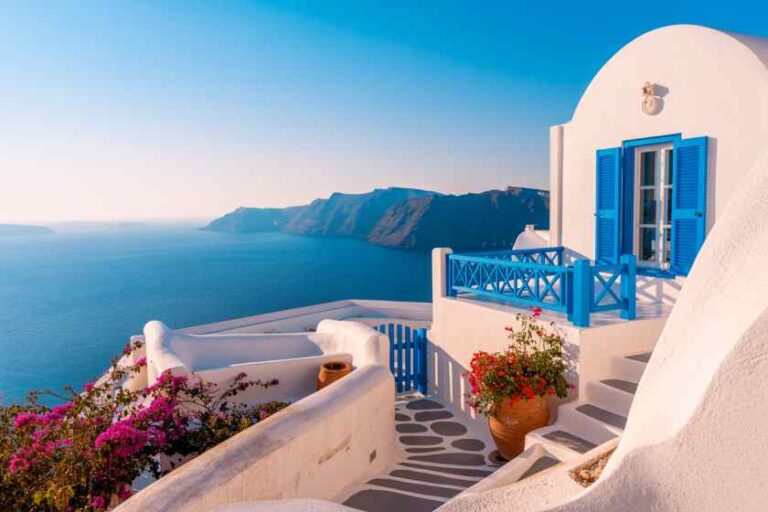 Santorini-style white house with blue shutters and balcony overlooking the blue Aegean Sea on a cliffside terrace.