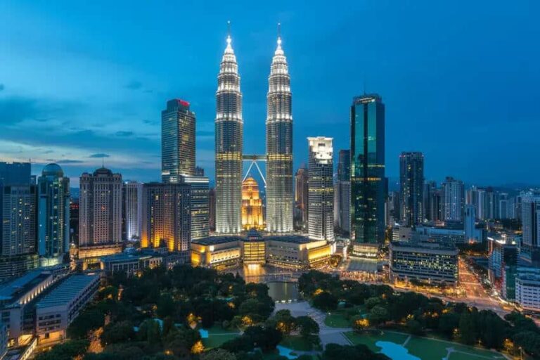 KL city skyline at dusk featuring the illuminated Petronas Twin Towers and surrounding skyscrapers with a central fountain plaza and green park in the foreground