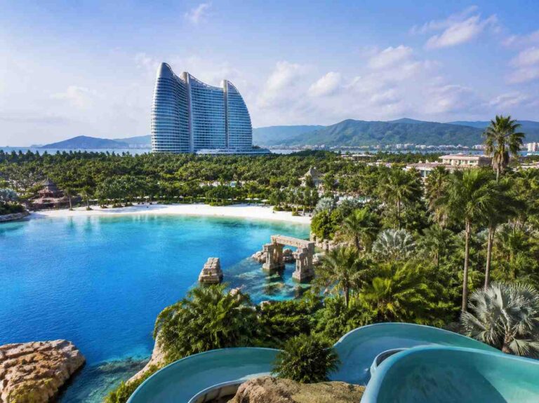 Turquoise lagoon with palm trees, a winding water slide in the foreground, and a large curved modern hotel in the distance.