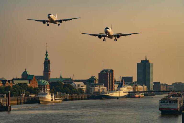 Two airplanes approaching to land over a riverfront city with boats and a sunset skyline.
