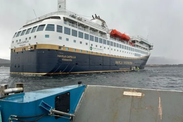 Large white and dark blue passenger ferry named Havila Capella seen from a small blue boat, approaching a dock on choppy water.