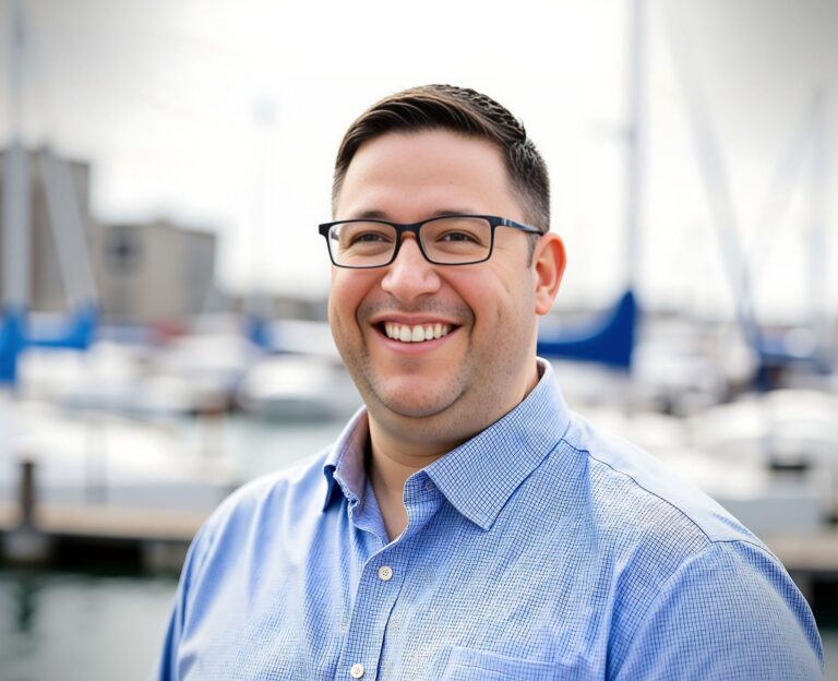 Close-up of a smiling man wearing black glasses and a light blue checkered shirt outdoors.