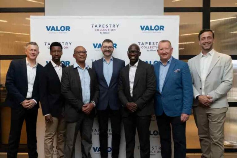 Seven men in business attire pose for a group photo in front of a backdrop displaying Valor and Tapestry Collection logos at a corporate event.