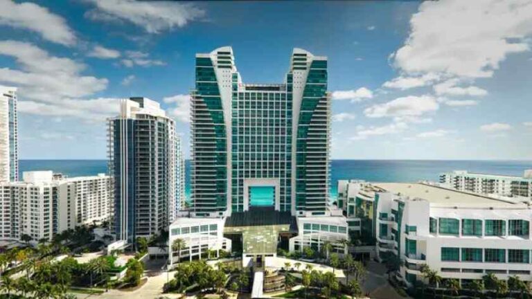 Modern beachfront hotel complex with two tall glass towers and a central entrance, facing the sea on a sunny day.