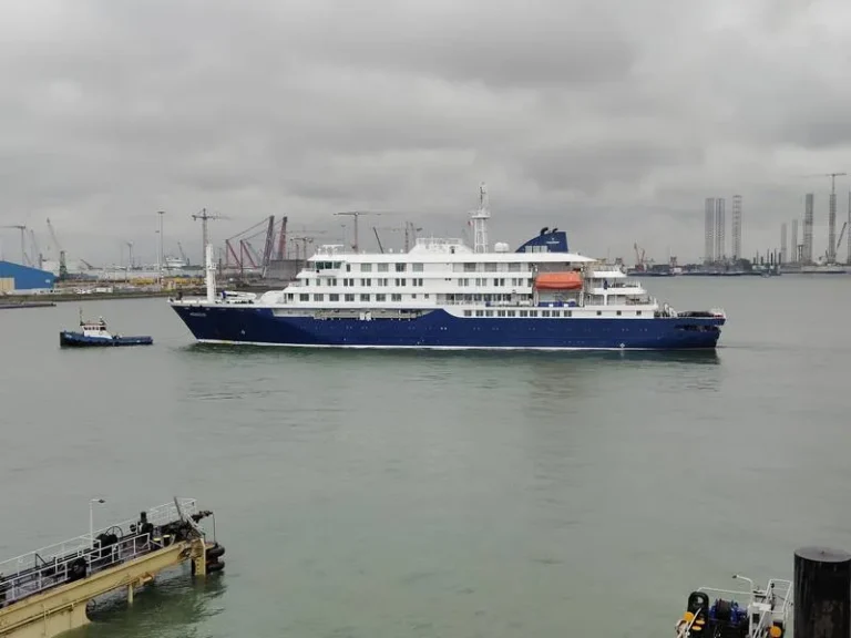 Large blue-and-white cruise ship in a calm harbor, with a small tugboat nearby and an industrial port with cranes in the background under cloudy skies.