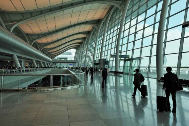 Interior of a modern airport terminal with a curved ceiling, large glass walls, and travelers with luggage on a reflective floor,