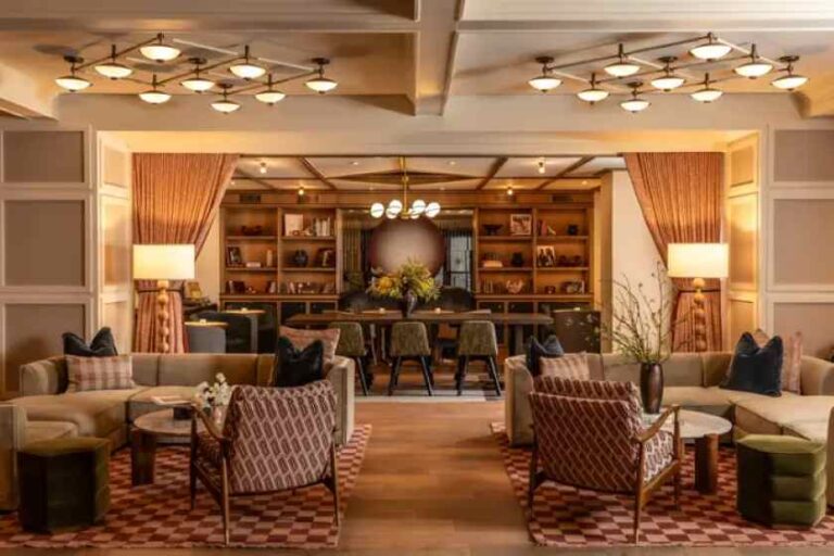 Hotel lounge with beige sofas, patterned chairs, and a back bar area; warm, ambient lighting and bookshelves nearby.