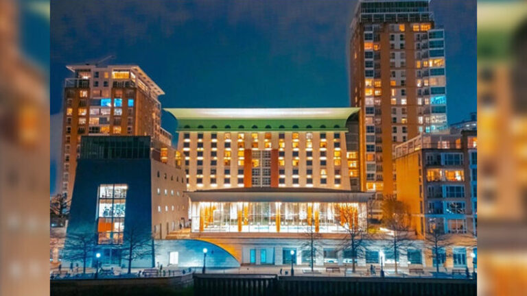 Nighttime view of a modern, multi-story building with a glowing glass entrance, surrounded by tall illuminated towers.