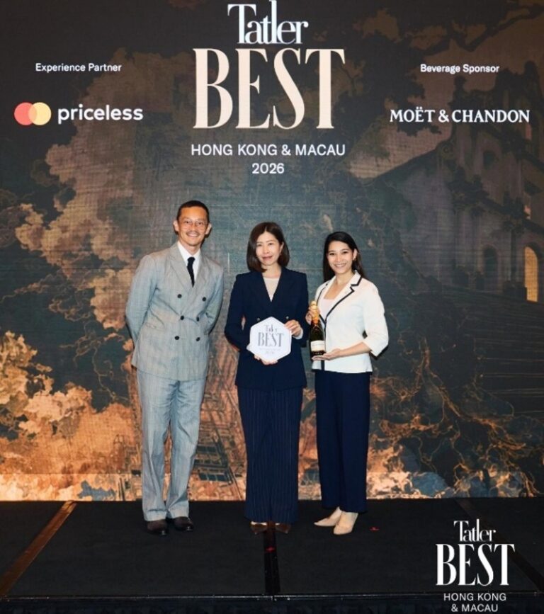 Three professionals stand on a stage at a Tatler Best Hong Kong & Macau 2026 event; the woman in the center holds a circular award while another woman holds a bottle of champagne, all against a backdrop with event branding.