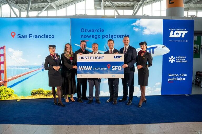 Six people in formal attire pose with a large 'First Flight' LOT banner at a travel event, in front of a San Francisco-themed backdrop.