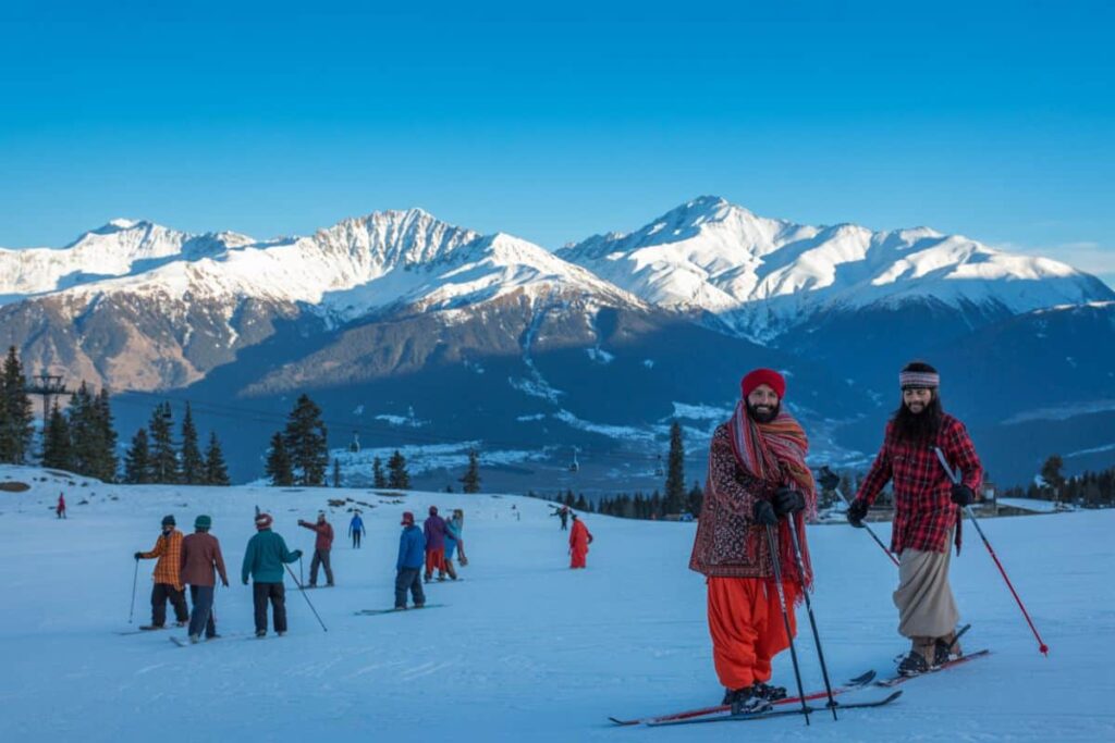 Group of people skiing on a snowy slope with a snow-capped mountain range under a clear blue sky; two men in colorful traditional clothing in the foreground.