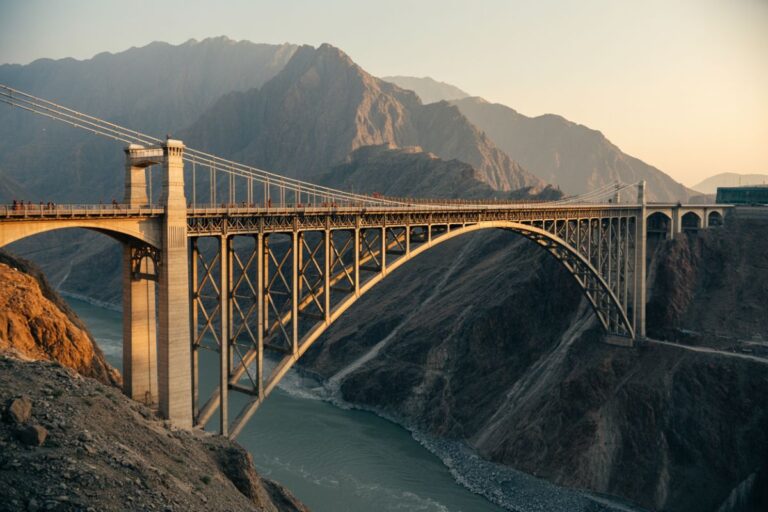 Suspension bridge spanning a deep canyon at sunset, with a river below and rugged mountains in the distance.