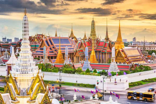 Sunset over a Thai temple complex with gilded spires, colorful roofs, and white surrounding walls in Bangkok-style architecture