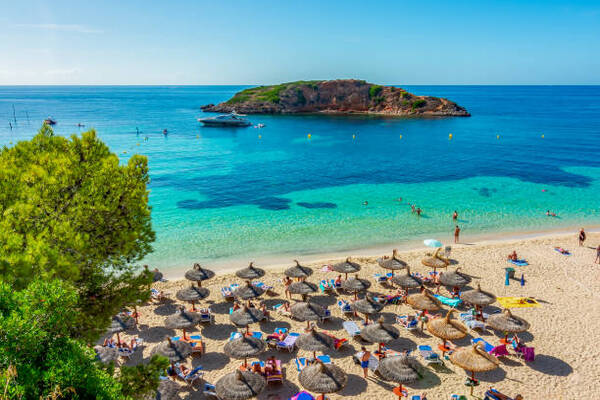 Busy beach with rows of straw umbrellas, sunbathers, and turquoise sea with a small island in the distance.