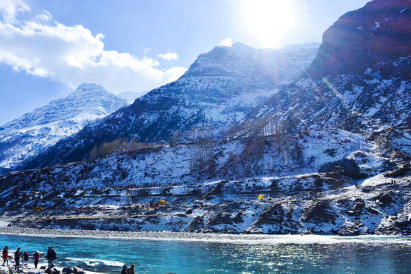 Snowy mountain range rises above a bright blue lake, with a group of people walking along the rocky, icy shore in sunlight