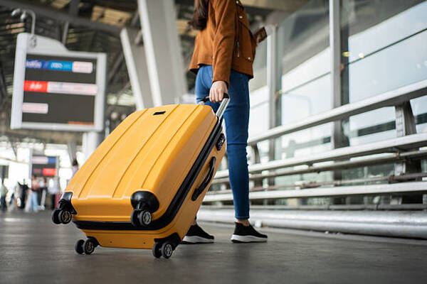 Person wearing a brown jacket and blue jeans pulls a yellow rolling suitcase through an airport terminal.