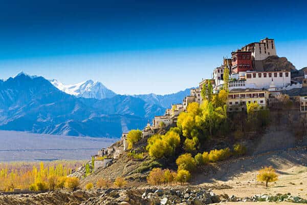 Hillside village of white buildings stacked on a slope, with yellow trees and distant snow‑capped mountains under a blue sky.