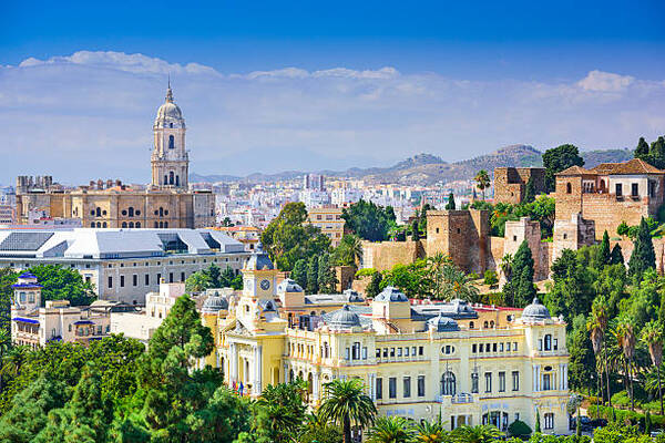 Panoramic view of a Mediterranean city with historic beige-and-yellow buildings, a tall church bell tower, fortress walls on a hill, palm trees, and distant hills under a blue sky