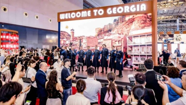 Choir in blue uniforms performing on a stage under a 'Welcome to Georgia' sign at an expo, with a crowd filming and watching nearby.
