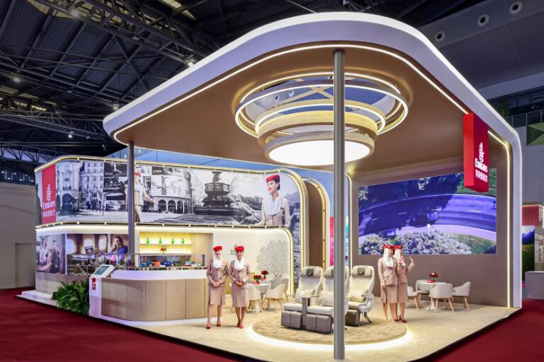 Emirates trade show booth with curved white-and-gold architecture, a reception desk, seating area, red-hat staff, and large display wall on a red carpeted floor.