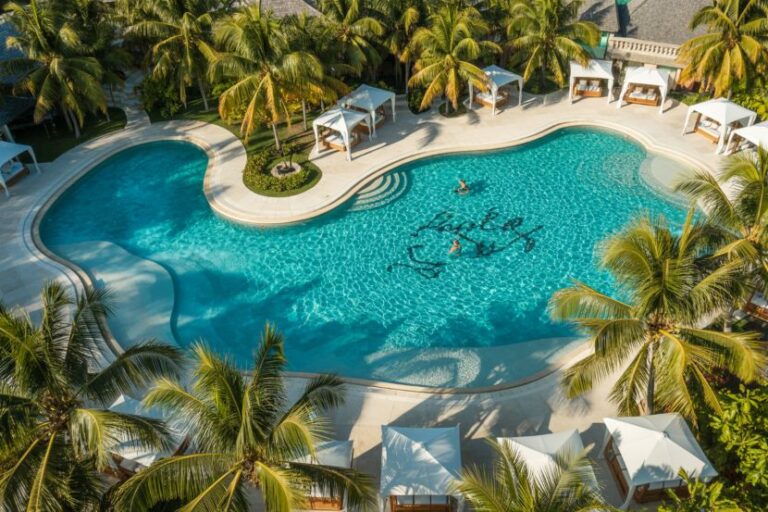 Aerial view of a resort pool surrounded by palm trees, cabanas, and lounge areas with turquoise water and sunlit surroundings.