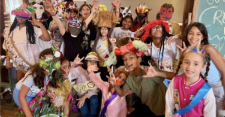 Group of children in colorful costumes posing for a photo at a costume party in a classroom setting.