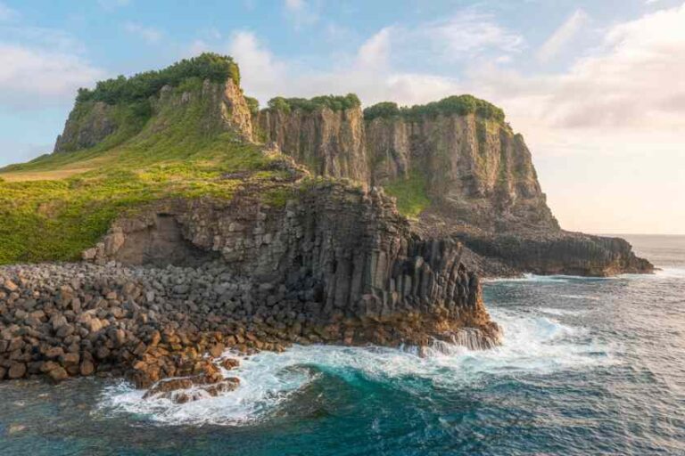 Cliffside rocky coastline with green vegetation atop columnar rocks meeting the sea.