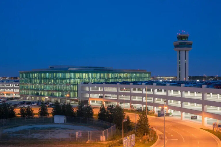 Airport terminal with glass facade and control tower at dusk, cars on a roadway in the foreground